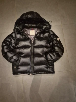 Svart dunjacka från Moncler - Snygg svart dunjacka från Moncler med glansig finish och klassisk logga på ärmen. Jackan har huva, dragkedja framtill och praktiska fickor. Perfekt för kalla vinterdagar och riktigt trendig look.