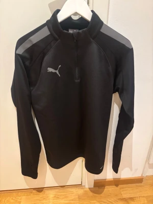 Svart träningströja från Puma - Svart långärmad träningströja från Puma med halv dragkedja och gråa detaljer på axlarna. Tröjan har en hög krage och är tillverkad i ett mjukt, stretchigt material som passar perfekt för sport och träning. Puma-logga i grått på bröstet.