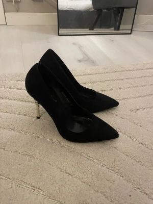 Svarta pumps med guldfärgad klack - Snygga svarta pumps med spetsig tå och guldfärgad smal klack. Skorna har en klassisk siluett, perfekta för dig som vill ha en elegant och trendig look.