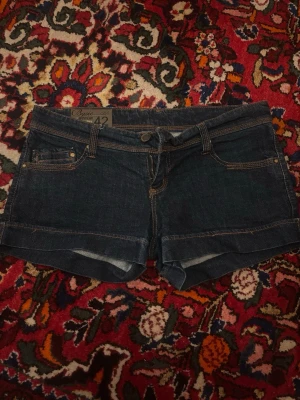 Mörkblå jeansshorts low waist - Snygga mörkblå jeansshorts från strandivarius basic med klassisk femficksdesign, synliga sömmar och uppvikta benslut. Shortsen har bälteshällor och detaljerade bakfickor med kontrastsömmar. Perfekta för varma dagar och enkel att matcha med allt.