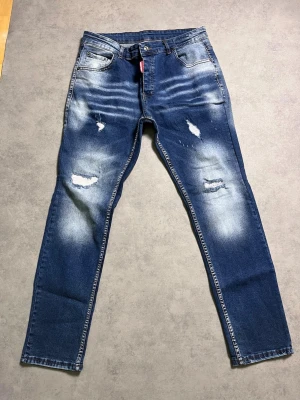 Dsquared2 jeans - Säljer ett par feta  jeans med slitningar och fades  på både lår och knän. Jeansen har klassisk femficksmodell, normalhög midja och tydliga tvättade partier för en trendig look. Materialet är denim i bomull med lite stretch. Sitter mer som w34 än w38 för att måttet ä 43cm tvärs över midjan