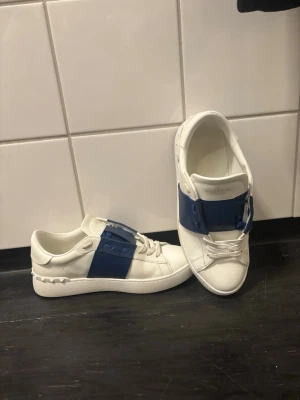 Mörk blåa valention skor  - Snygga vita sneakers från Valentino Garavani med bred blå rem över mitten och klassiska nitar på hälen. Skorna är väldigt snygg o trendiga. Skor har knappast används pga för små. Ny pris är runt 5000 