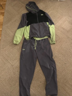 Under Armour träningsset hoodie & byxor - Snyggt träningsset från Under Armour med hoodie och byxor i grått, svart och ljusgrönt. Setet har dragsko i midjan och på luvan, samt logga på både jacka och byxa. Perfekt för gymmet eller löprundan.