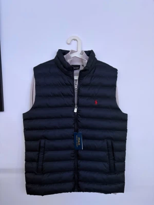 Svart dunväst från Ralph Lauren - Snygg svart dunväst från Ralph Lauren med diskret röd logga på bröstet och broderad text på ryggen. Västen har ståkrage, dragkedja framtill och två fickor med dragkedja. Perfekt för lager-på-lager och riktigt stilren look.