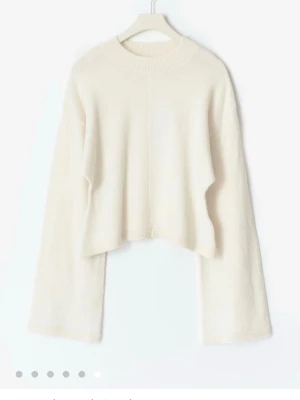 Vit croppad stickad tröja - Trendig vit stickad tröja med croppad passform och vida ärmar. Tröjan har en enkel design med rund halsringning och diskret söm framtill. Perfekt för att styla med höga jeans eller kjol för en clean look. Har ett litet håll vid axel som var där när jag köpte den men inget som syns stort, annars helt ny🙏🏼🥰