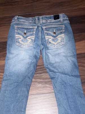 Blå bootcut jeans med broderade fickor - Snygga blå bootcut jeans med broderade bakfickor och knappar. Jeansen har ljusa slitningar och klassisk femficksdesign. Materialet är denim och passformen är något utsvängd nedtill. Perfekt för dig som gillar detaljer och en avslappnad look.