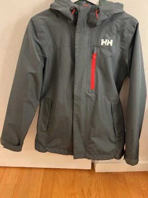 Grön Helly Hansen vindjacka med huva - Snygg grå vindjacka från Helly Hansen med huva och röda detaljer på dragkedjor och snören. Jackan har Helly Tech-tryck på ärmen, tre fickor och HH-logga på bröstet. Materialet är vattenavvisande och perfekt för blåsiga dagar.