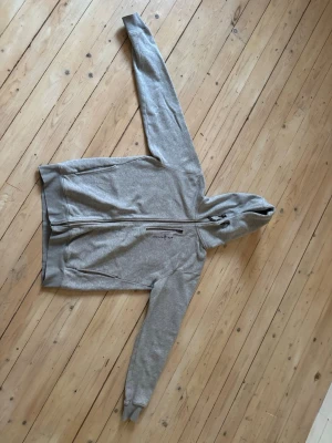 Grå zip hoodie med huva - Snygg grå zip hoodie från Sail Racing med dragkedja framtill och två fickor. Klassisk design med huva och ribbade muddar vid ärmslut och nederkant. Perfekt för en avslappnad och alldaglig stil. Tröjan är använd någon enstaka gång och är i perfekt skick!