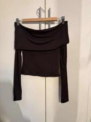 Brun offshoulder topp från H&M - Trendig svart offshoulder topp från H&M med långa ärmar och croppad passform. Tillverkad i mjukt stretchigt material som sitter snyggt på kroppen. Perfekt att styla med höga jeans eller kjol för en cool look.