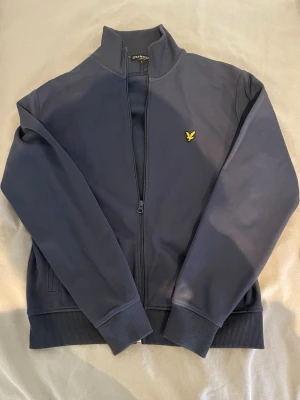 Lyle & Scott mörkblå zip (äkta) - Snygg mörkblå hoodie från Lyle & Scott med dragkedja framtill och ribbade muddar. Klassisk gul logga på bröstet och två fickor. Tillverkad i mjukt material som känns skönt mot huden. Perfekt för en avslappnad och stilren look. Storlek M/L. 
