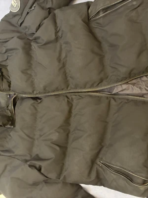 Svart pufferjacka från Supply & Demand - Snygg svart pufferjacka från Supply & Demand med patch på ärmen och dragkedjor i metall. Jackan har vadderad design, hög krage och praktiska fickor med dragkedja. Perfekt för kalla vinterdagar och ger en cool streetwear-look.