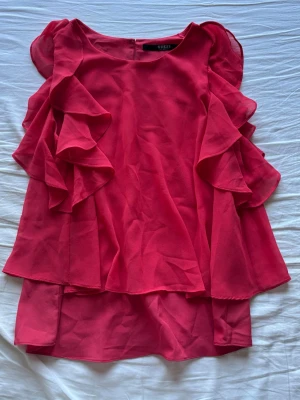 Rosa/röd volangblus från Guess - Säljer en snygg rosa/röd blus från Guess med volangdetaljer längs sidorna. 