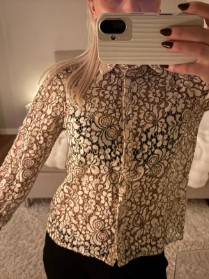 Beige spetsblus med blommönster - Säljer en beige spetsblus med detaljerat blommönster och knäppning framtill. Blusen har lång ärm och klassisk krage, perfekt för dig som gillar en romantisk och trendig stil. Materialet är genomskinligt spets som ger en cool och elegant vibe.