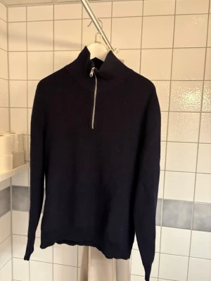 Mörkblå stickad tröja med halvzip - Mörkblå stickad tröja med hög krage och halv dragkedja framtill. Tröjan har långa ärmar och är ribbstickad vid kragen. Perfekt för lager-på-lager och enkel att matcha med jeans eller byxor. Passar small/medium. Använd max 5 ggr 
