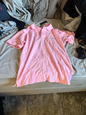 Rosa pikétröja från Ralph Lauren - Snygg rosa pikétröja från Ralph Lauren med klassisk krage och korta ärmar. Tröjan har den ikoniska broderade loggan på bröstet och är tillverkad i mjuk bomull. Perfekt för en avslappnad och stilren look.