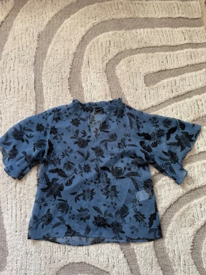 Blå blommig blus med volangkrage - Snygg blå blus med svart blommönster och volangdetalj vid kragen. Blusen har korta, vida ärmar och en liten knappöppning framtill. Tunt, lätt och transparent material som ger en luftig känsla. 