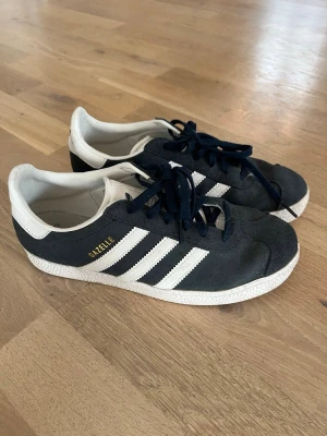 Adidas Gazelle marinblå sneakers - Klassiska Adidas Gazelle sneakers i marinblå mocka med vita detaljer och de ikoniska tre ränderna på sidan. Låg modell med vit sula och guldfärgad Gazelle-text. Sköna och stilrena skor som passar till det mesta.