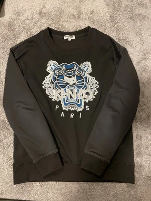 Svart sweatshirt med tiger från Kenzo - Svart sweatshirt från Kenzo med ikoniskt broderat tigerhuvud i blått, vitt och grått på bröstet samt texten 'KENZO PARIS'. Tröjan har rund halsringning, ribbade muddar och är tillverkad i mjukt bomullsmaterial. Perfekt för en trendig och avslappnad stil.