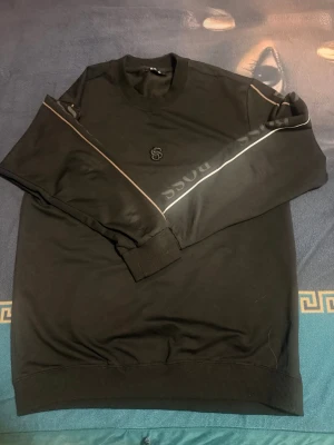 Svart stickad tröja från BOSS - Svart stickad tröja från BOSS med rund halsringning och ribbade muddar. Tröjan har en diskret BOSS-logga på bröstet och en dekorativ rand längs ärmarna med BOSS-tryck. Perfekt för en stilren och modern look.