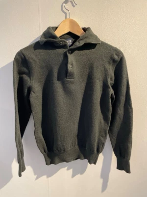 Mörkgrön Kashmir tröja med krage - Mysig mörkgrön stickad tröja från Woolpower med klassisk krage och knäppning framtill. Tröjan har långa ärmar, ribbade muddar och är tillverkad i ull vilket gör den varm och skön. Perfekt för kyliga dagar och enkel att matcha. 