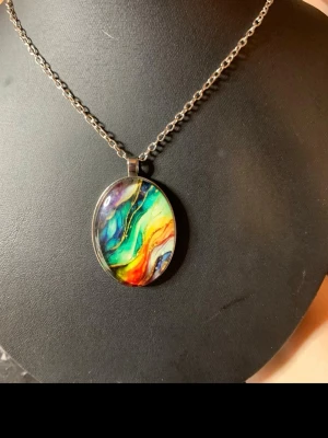Färgglatt halsband med abstrakt hänge - Unikt halsband med ett ovalt hänge i regnbågens färger och abstrakt mönster. Hänget har en glansig yta och sitter på en silverfärgad kedja. Perfekt för dig som vill sticka ut med färg och kreativ stil.