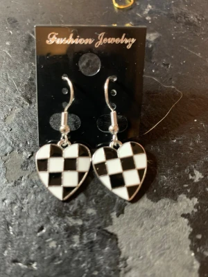 Hjärtformade örhängen med schackmönster - Säljer ett par hjärtformade örhängen i svartvitt schackmönster. De har silverfärgade krokar och en cool retrostil som sticker ut. Perfekta för dig som gillar statement-accessoarer och vill addera lite edge till din look.