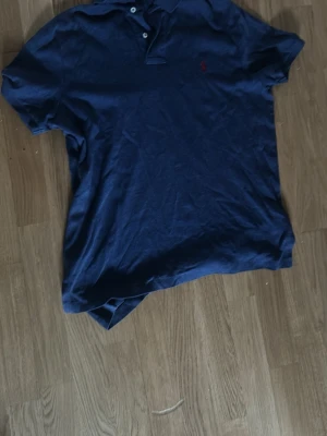 Marinblå pikétröja från Polo Ralph Lauren - Klassisk marinblå pikétröja från Polo Ralph Lauren med krage och knappar framtill. Tröjan har en liten röd logga broderad på bröstet och är tillverkad i mjuk bomull. Perfekt för en stilren och avslappnad look.