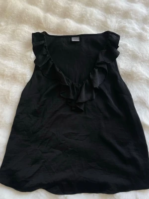 Svart linnetopp med volang - Snygg svart ärmlös linnetopp med djup v-ringning och volangdetaljer längs urringningen. Perfekt att styla med jeans eller kjol för en trendig look. Luftigt material som passar till varma dagar.