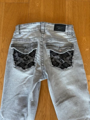 Ljusgrå jeans med spetsdetalj - Säljer ett par ljusgrå bootcut jeans med snygga svarta spetsdetaljer på bakfickorna. Spets detaljen har jag sytt dit själv och de är lite slarvigt gjort. Jeansen är i jättebra skick och knappt använda, pris kan diskuteras.
