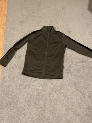 Olivgrön träningsjacka från Under Armour - Snygg olivgrön träningsjacka från Under Armour med hel dragkedja och svarta detaljer längs ärmarna. Jackan har hög krage och är tillverkad i ett lätt och stretchigt material som passar perfekt till träning eller uppvärmning. Passar både S och M