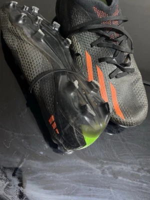 Adidas svarta fotbollsskor med orange detaljer - Snygga fotbollsskor från Adidas i svart med orangea ränder och detaljer. Skorna har en strömlinjeformad design, snörning och ovandel i mesh-liknande syntetmaterial. Yttersulan har fasta dobbar för spel på gräs. Perfekta för dig som vill ha stil och fart på planen.