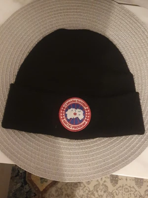 Svart mössa från Canada Goose - Säljer en svart ribbstickad mössa från Canada Goose med klassisk uppvikt kant och det ikoniska runda märket framtill. Perfekt för kalla dagar och riktigt snygg till alla ytterkläder. Materialet känns mjukt och värmande.