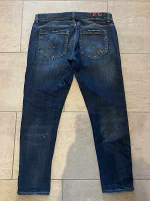 Mörkblå dondup jeans - Säljer ett par mörkblå dondup  slim fit jeans med lätt slitna detaljer och diskreta färgstänk. Klassisk femficksmodell med knappgylf och smal passform. Perfekta för en avslappnad och trendig look. Jeans passar någon som är runt.  170- 175