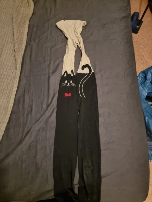 Tights med kattmotiv och rosett - Säljer ett par tights med beige överdel och svart nedtill. På låren finns ett gulligt kattmotiv med vita morrhår, öron och svans samt en liten röd rosett. Perfekta för dig som gillar söta detaljer och djurmotiv.