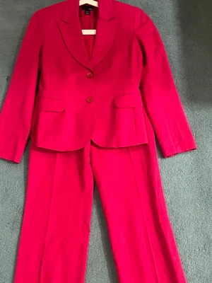 Kostym i stark rosa med kavaj och byxa - Snygg kostym i stark rosa färg med klassisk kavaj och matchande byxa. Kavajen har två knappar framtill, breda slag och fickor med lock. Byxorna är raka och har en stilren passform. Perfekt för dig som vill sticka ut med färg.