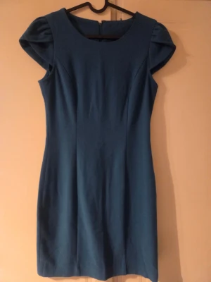 Blå kortärmad klänning med puffärm - Snygg blå klänning med rund halsringning och korta puffärmar. Klänningen har en figurnära passform och dragkedja i ryggen. Perfekt för dig som gillar enkel men elegant stil. I petroleumgrön färg. 