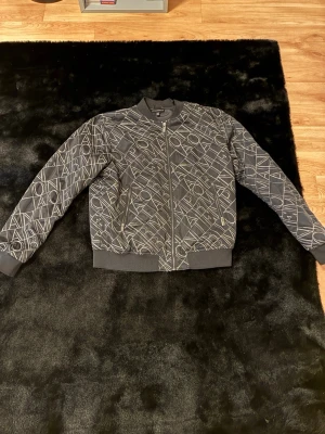 Svart bomberjacka med grafiskt mönster - Snygg svart bomberjacka från Emporio Armani med unikt grafiskt mönster i vitt över hela jackan. Jackan har ribbade muddar, dragkedja framtill och mjukt foder. Perfekt för dig som vill sticka ut med en stilren och modern look.