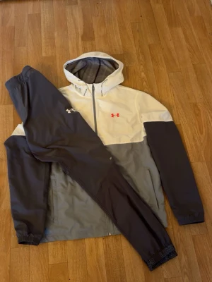 Under Armour träningsset med huva - Snyggt träningsset från Under Armour med jacka och byxor. Jackan har huva, dragkedja och är vit och grå med svarta ärmar samt röd logga på bröstet. Byxorna är svarta med logga på låret. Perfekt för träning eller chill.