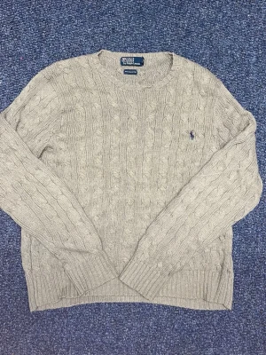 Kabelstickad tröja Ralph Lauren - Kabelstickad grå tröja från Polo Ralph Lauren med rund halsringning och broderad logga på bröstet. Tröjan har långa ärmar och ribbade muddar vid ärmslut och nederkant. Perfekt för en stilren och klassisk look. 100% äkta