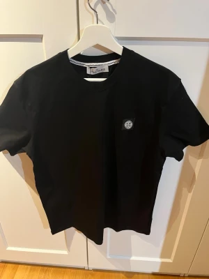 Svart t-shirt från Stone Island - Svart t-shirt från Stone Island med klassisk rund hals och korta ärmar. T-shirten har en liten patch med Stone Island-loggan på bröstet. Enkel och stilren design i mjuk bomull, perfekt till jeans eller shorts. Storlek M men sitter som L