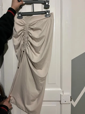 Beige omlottkjol med snörning - Trendig beige kjol med snygg snörning och omlottdetalj framtill. Kjolen har en draperad effekt som ger en cool och modern look och slits. Perfekt för dig som vill sticka ut med en unik stil. Materialet känns mjukt och följsamt.