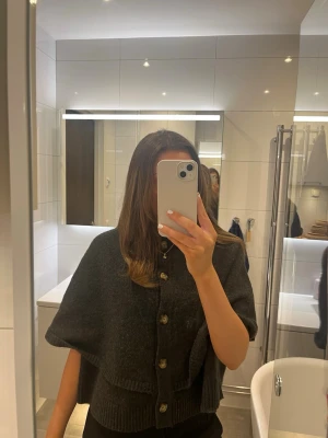 Mörkgrå cape-kofta med knappar - En snygg mörkgrå cape-kofta med stora knappar framtill och ribbade detaljer vid nederkant. Koftan har en lös och boxig passform som ger en cool look. Perfekt att slänga över en t-shirt eller topp för extra värme. Materialet ser ut att vara mjukt och stickat. Vid snabb affär går de bra att diskutera prissänkning!