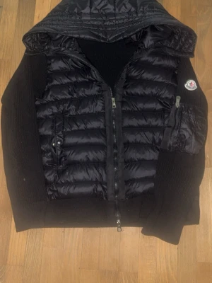 Moncler cardigan  - Snygg svart hoodie från Moncler med vadderad dunpanel framtill och på huvan. Tröjan har dragkedja, ficka med lock på bröstet och Moncler-logga på ärmen. Perfekt för dig som gillar streetwear och exklusiva märken.