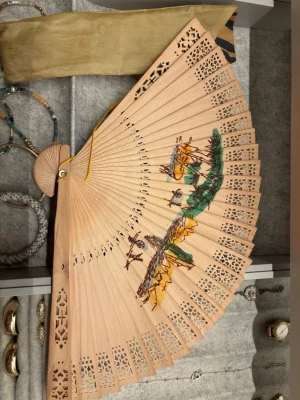 Solfjäder  - Snygg handfläkt i ljust trä med utskurna detaljer och handmålat landskapsmotiv i grönt, gult och brunt. Fläkten kommer med ett guldfärgat tygfodral. Perfekt accessoar för att ge din stil en unik touch.