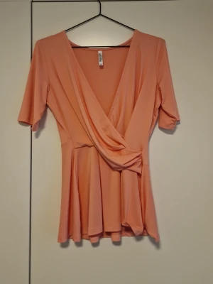 Rosafärgad omlottopp  - Trendig rosafärgad omlottopp i midjan och v-ringad hals. Toppen har korta ärmar och ett lätt utsvängt fall nedtill. Perfekt för dig som gillar en feminin och bekväm stil.