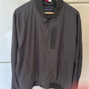 Grå bomberjacka från Tommy Hilfiger - Snygg grå bomberjacka från Tommy Hilfiger med klassisk ribbad krage och muddar. Jackan har dragkedja framtill, praktiska fickor och en stilren look. Perfekt för dig som gillar enkel och clean stil med en touch av street.