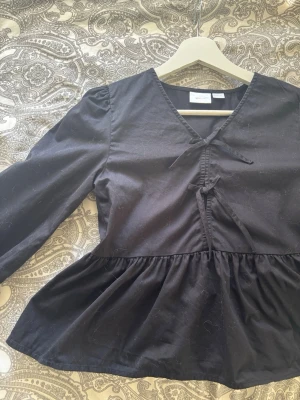 Svart blus med volang och knytning - Snygg svart blus från Gina Tricot med volang nertill och knytning framtill vid halsen. Blusen har v-ringning och långa ärmar. 