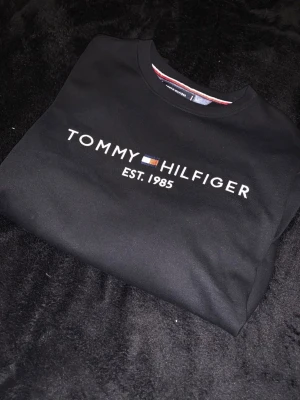 Svart Tommy Hilfiger hoodie - Snygg svart hoodie från Tommy Hilfiger med klassisk logga och texten 'EST. 1985' broderat på bröstet. Tröjan har rund halsringning och är tillverkad i mjukt material som känns skönt mot huden. Perfekt för en stilren och avslappnad look.