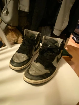Nike Air Jordan 1 svart/grå high - Säljer ett par Nike Air Jordan 1 high sneakers i svart och grått. Klassisk siluett med högt skaft, svart swoosh och vingar-logga på sidan. Ovandel i läder och textil, vita sulor och svarta skosnören. Perfekta för dig som gillar streetwear och retrostil.