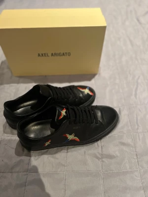 Svarta Axel Arigato skor - Snygga svarta sneakers från Axel Arigato i läder med färgglada broderade fåglar på sidan. Klassisk låg modell med svarta skosnören och diskret logga på sidan. Perfekta för dig som vill ha en clean look med en twist. Skorna är andvända typ två gånger pris kan diskuteras vid snabb affär box ingår !